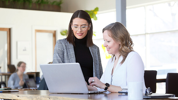 two women one standing one sitting looking at a laptop in an office environment (photo)