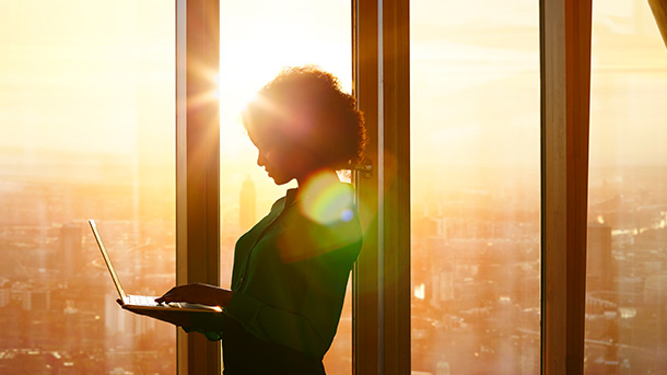 Woman in a glass office with a sunset behind her looking at a laptop (photo)