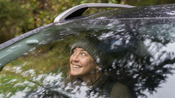 Happy woman sitting in her car in a forest looking up at the trees. (photo)