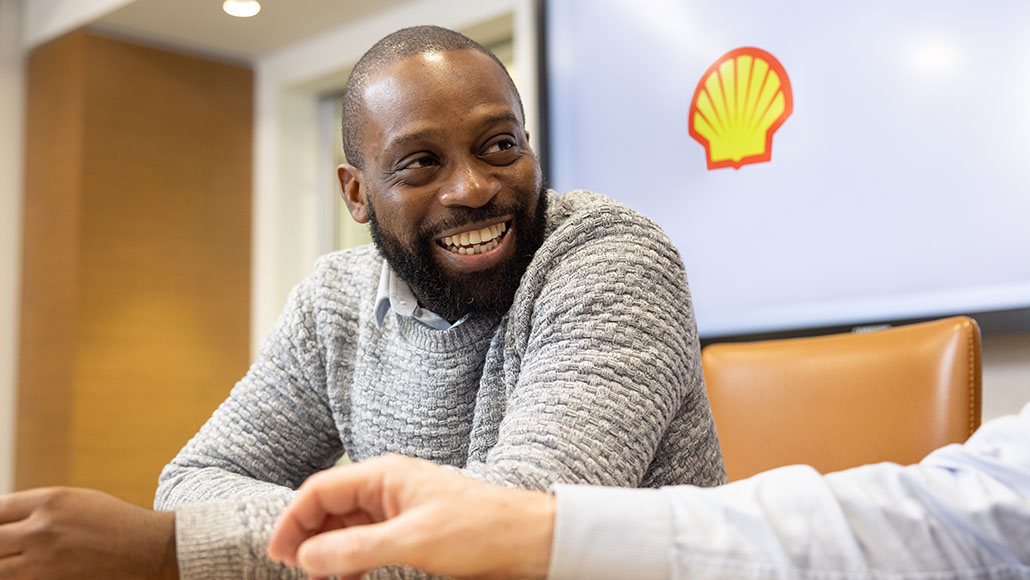 Man smiling and sitting in a Shell office with the Shell pecten on a wall in the background talking to another person (photo)