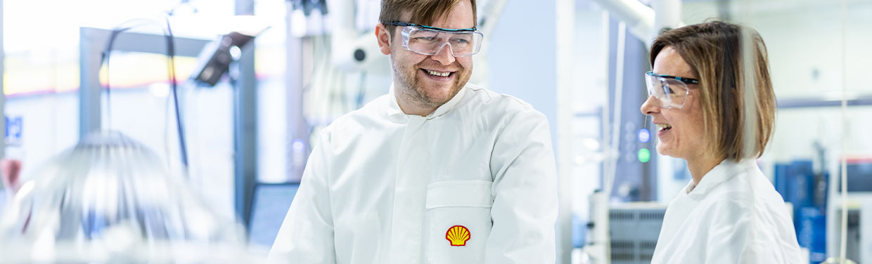 Two people in a laboratory wearing safety goggles and Shell lab coats smiling at each other (photo)