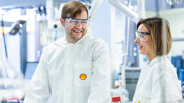 Two people in a laboratory wearing safety goggles and Shell lab coats smiling at each other