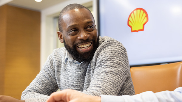 Man smiling and sitting in a lounge with the Shell logo on a wall in the background talking to another person