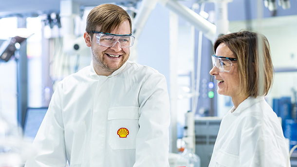 Two people in a laboratory wearing safety goggles and Shell lab coats smiling at each other