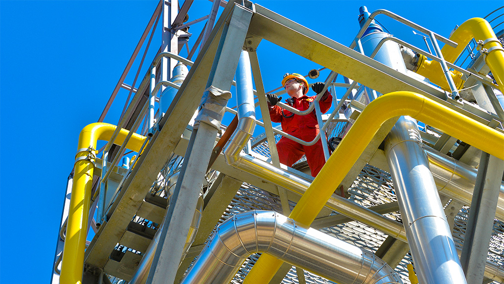 Shell employee standing on a platform at a refinery showing yellow pipes against a blue sky