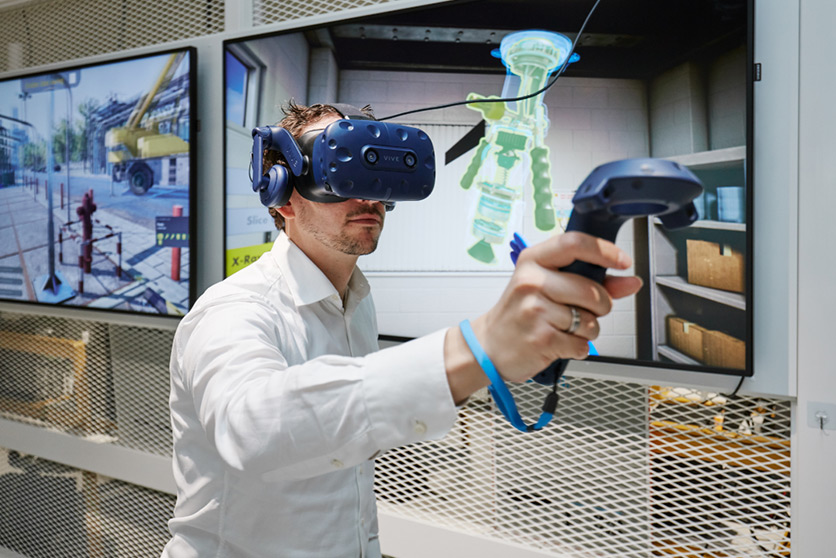 An employee testing a virtual reality headset and controls in front of large screens showing 3D images (photo)