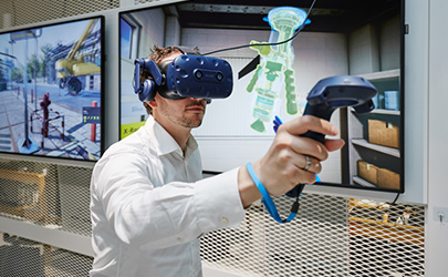 An employee testing a virtual reality headset and controls in front of large screens showing 3D images (photo)