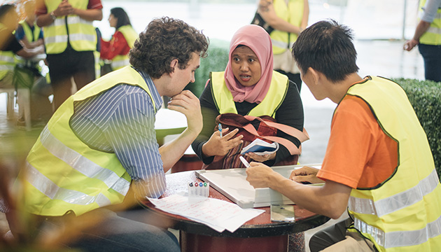 Three entrepreneurs from the UK, Malaysia and Indonesia who all wear reflective vests discussing around a small table. (photo)