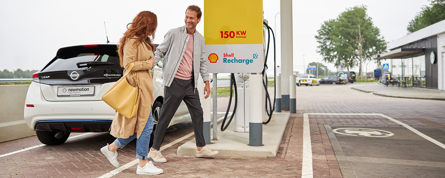 A woman and a man walking at a service station in the UK, as their electric car charges at the charging station. (photo)