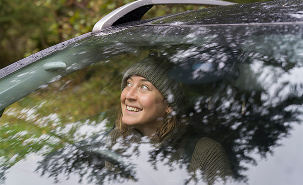 Smiling woman wearing a cap looking out of the front window of a car surrounded by trees (photo)