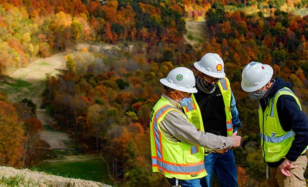 Three men wearing safety equipment and masks discussing safety topics near a pipeline which crosses a wood in the background (photo)