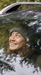 Smiling woman wearing a cap looking out of the front window of a car surrounded by trees (photo)