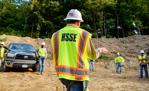Employee wearing hard hat and hi-vis vest printed with HSSE leading a safety briefing outside (photo)