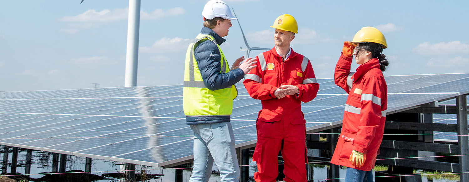two men and a woman wearing PPE standing together talking next to a solar panel with wind turbine in the background (photo)