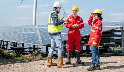 two men and a woman wearing PPE standing together talking next to a solar panel with wind turbine in the background (photo)