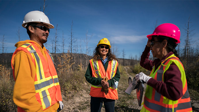 Three people in safety gear are conversing in a field with sparse trees in the background (photo)