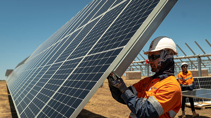 Workers are installing solar panels in a sunny field. The person in the foreground is wearing a white helmet and orange safety gear, adjusting a solar panel (photo)