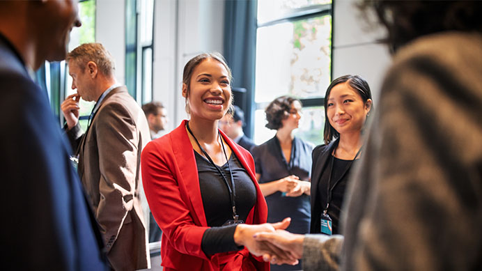 Two smiling women are shaking hands at a professional event, with other attendees in the background (photo)