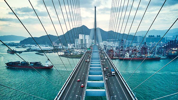 Image of a suspension road bridge crossing in Hong Kong with containers freight ship passing underneath and urban cityscape in the background. (photo)